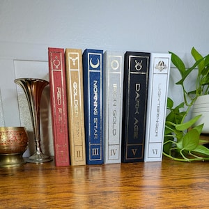 May include: A collection of decorative books with gold lettering and accents, arranged on a wooden surface. The books are in various colors, including red, gold, blue, and black. A brass vase and a decorative bowl are also present.