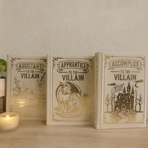 May include: Three hardback books with gold foil lettering and illustrations. The titles read "Assistant to the Villain," "Apprentice to the Villain," and "Accomplice to the Villain." The books are cream-coloured and feature fantasy-themed artwork.