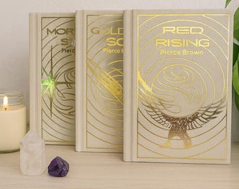 May include: Three hardcover books with gold foil lettering and designs. The book titles are "Morning Star", "Golden Son", and "Red Rising" by Pierce Brown. A lit candle, quartz crystal, and amethyst are in the foreground.