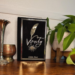 May include: A hardcover book titled "Verity" by Colleen Hoover. The book has a black cover with gold foil accents, including a feather quill and the title in elegant script. The book is displayed on a wooden surface.