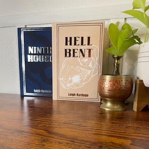 Könnte beinhalten: Zwei Taschenbücher von Leigh Bardugo, "Ninth House" und "Hell Bent", liegen auf einer Holzoberfläche. Die Buchcover sind blau und braun mit weißem Text und Illustrationen.