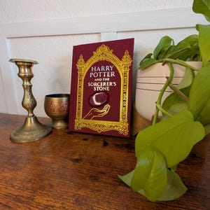 May include: A maroon hardcover book titled "Harry Potter and the Sorcerer's Stone" with gold lettering and design. The book is propped up on a wooden surface next to a brass candlestick, a small decorative cup, and a potted plant.
