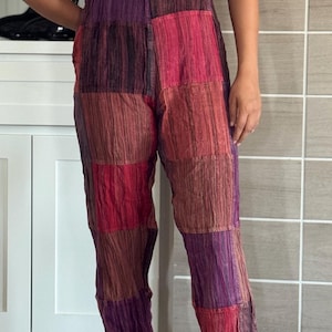 May include: Multi-colored patchwork overalls in shades of purple, red, and brown. The overalls have a bib pocket and adjustable straps. The model is wearing a black t-shirt and white sneakers.