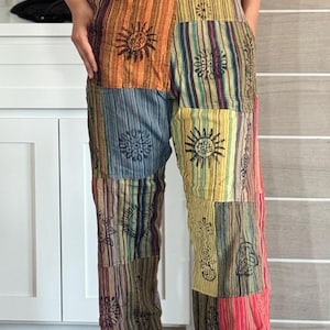 May include: Patchwork overalls in a variety of colors including orange, blue, and red. The overalls feature a striped pattern with sun and floral designs. The garment has adjustable straps and pockets. The person is wearing white sneakers.