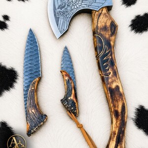 Puede incluir: Un conjunto que consta de un hacha grande y dos cuchillos con hojas de metal oscuro texturizado y mangos de madera quemada curvados. La cabeza del hacha presenta una figura alada grabada. Un logotipo dorado con el texto AYZO CREEK KNIVES es visible.
