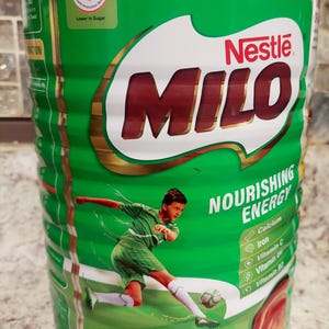 Pode incluir: Uma lata verde e dourada de pó Milo com uma imagem de um menino jogando futebol. A lata diz "Milo Powder" e "Nourishing Energy" e tem um logotipo da Nestlé. A lata é de 1,8 kg e tem menos açúcar.