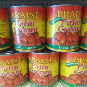 May include: A stack of canned goods labeled "PRAISE Palm Cream." The cans have a red and yellow label with an image of red palm fruits. The text "SAUCE GRAINE" is printed at the bottom of the label.