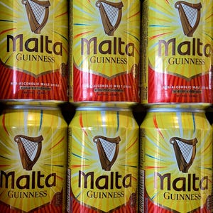 May include: Six yellow Malta Guinness cans are stacked. Each can features a brown harp logo and the words "Malta Guinness" in large letters. The cans also state "Non-Alcoholic Malt Drink" and have a red and orange design at the bottom.