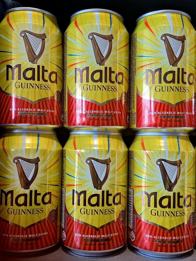 May include: Six yellow Malta Guinness cans are stacked. Each can features a brown harp logo and the words "Malta Guinness" in large letters. The cans also state "Non-Alcoholic Malt Drink" and have a red and orange design at the bottom.