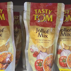 May include: Multiple pouches of "Tasty Tom Jollof Mix" are displayed. The packaging is red and gold, with images of tomatoes, onions, and a prepared Jollof dish. The text "Jollof Mix" is prominently featured.