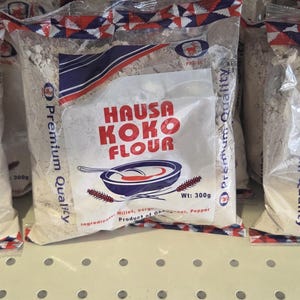Könnte beinhalten: Drei Beutel Hausa Koko Mehl werden auf einem Regal präsentiert. Jeder transparente Plastikbeutel zeigt den Produktnamen in Rot und Weiß sowie eine Schüsselgrafik. Die Beutel sind versiegelt und mit einem feinen, hellfarbigen Pulver gefüllt.