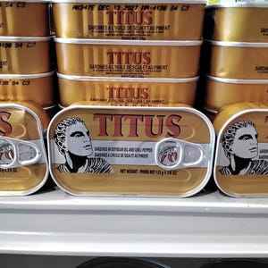 May include: A stack of canned sardines in soybean oil and chili pepper. The cans are yellow with a red and white label that says "TITUS" and the net weight is 125g (4.38 oz).