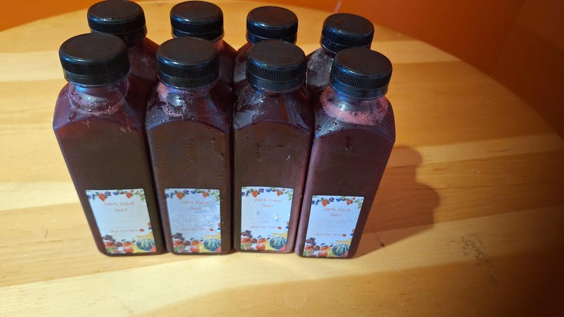 May include: Eight glass bottles filled with a dark red liquid. The bottles have black lids and labels with colorful fruit illustrations and the text "Berry Blush" on them.