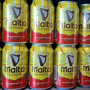 May include: A collection of yellow and red Malta Guinness cans. Each can features the Malta Guinness logo with a harp and the words "Malta Guinness" in bold text. The cans are arranged in neat rows.