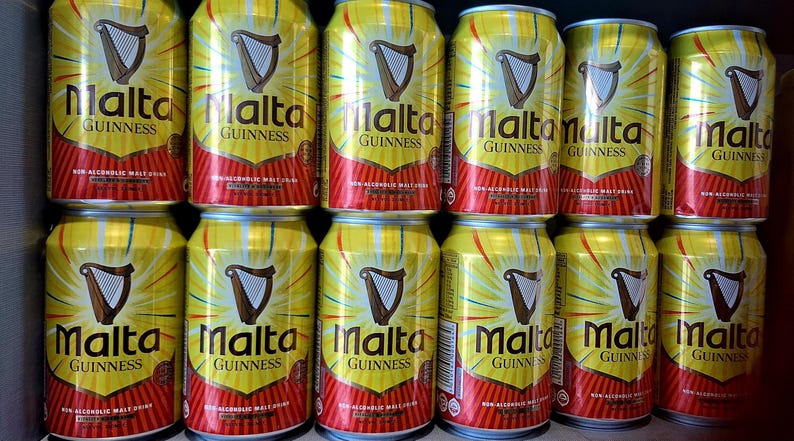 May include: A collection of yellow and red Malta Guinness cans. Each can features the Malta Guinness logo with a harp and the words "Malta Guinness" in bold text. The cans are arranged in neat rows.