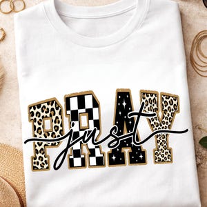 Könnte beinhalten: Weißes T-Shirt mit den Worten "PRAY just" in einem auffälligen Design. Die Buchstaben "P" und "Y" haben ein Leopardenmuster, während das "R" ein schwarz-weißes Schachbrettmuster ist. Der Buchstabe "A" ist schwarz mit weißen Sternen.