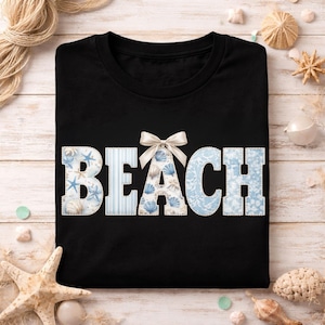 Puede incluir: Camiseta negra con la palabra "BEACH" en letras blancas, azules y con estampado de conchas marinas. Un lazo blanco está sobre la letra "A". La imagen incluye conchas marinas y otras decoraciones con temática de playa.