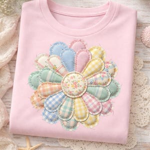 May include: A light pink t-shirt featuring a large, colorful flower design. The flower petals are made of various pastel-colored fabrics, including gingham and floral patterns, stitched together. The center of the flower has a floral pattern.