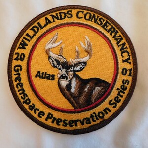 May include: Embroidered patch with a deer illustration. The patch is circular, featuring a deer head in the center, surrounded by text that reads "Wildlands Conservancy" and "Green Space Preservation Series". The patch has a brown border and a yellow background.