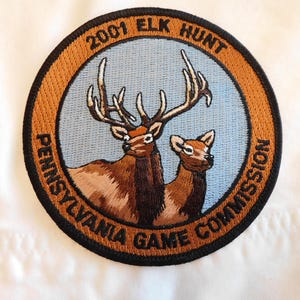 May include: Embroidered patch featuring a brown elk and a fawn against a light blue background. The patch is circular with the text "2001 ELK HUNT" and "PENNSYLVANIA GAME COMMISSION" in a brown and black border.