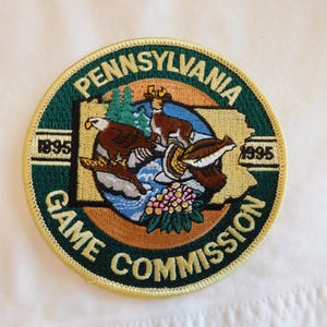 May include: Embroidered patch featuring the Pennsylvania Game Commission logo. The circular patch has a green border with the words "PENNSYLVANIA GAME COMMISSION" and the years "1895" and "1995". The center depicts an eagle, deer, and other wildlife.
