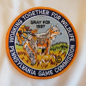 May include: Embroidered patch featuring a gray fox in a grassy field with fall foliage. The patch is circular with text that reads "Working Together for Wildlife" and "Pennsylvania Game Commission". The year "1997" is also displayed.