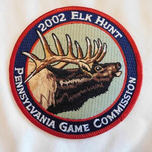 May include: Embroidered patch featuring a detailed elk with large antlers, set against a light green background. The patch is circular, with text reading "2002 ELK HUNT" and "PENNSYLVANIA GAME COMMISSION" in a blue and red border.