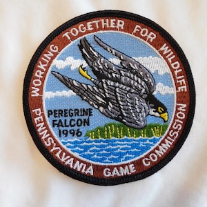 May include: Embroidered patch featuring a peregrine falcon in flight over water and land. The patch is circular with text that reads "Working Together for Wildlife" and "Pennsylvania Game Commission 1996". The falcon is gray, black, and white.