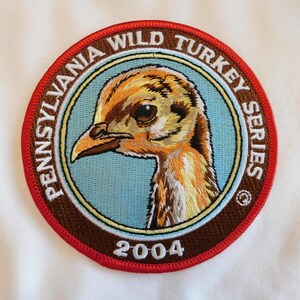May include: Embroidered patch featuring a detailed illustration of a wild turkey's head. The patch is circular, with text reading "PENNSYLVANIA WILD TURKEY SERIES" and the year "2004". The design includes shades of brown, orange, and blue.