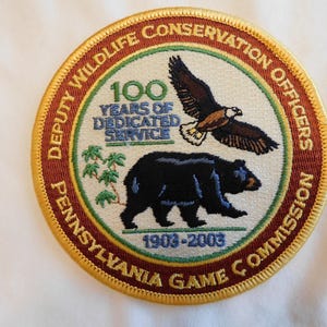 May include: Circular patch with a brown border and gold trim. The center features an eagle and a black bear, with text reading "100 Years of Dedicated Service" and "Pennsylvania Game Commission 1903-2003".