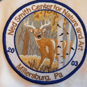 May include: Embroidered patch featuring a deer in a forest scene with birch trees and fall foliage. The patch is circular with a blue border and text that reads "Ned Smith Center for Nature and Art, Millersburg, Pa, 2003".