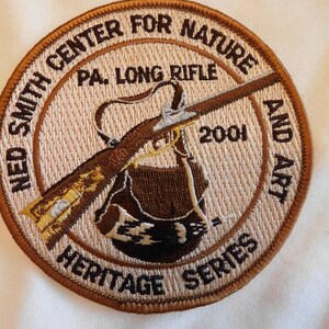 May include: Embroidered patch featuring a brown long rifle, a brown bag, and the text "NED SMITH CENTER FOR NATURE AND ART" and "PA. LONG RIFLE 2001 HERITAGE SERIES" in a circular design.