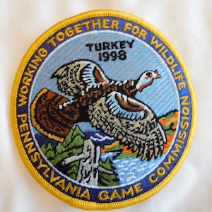 May include: Embroidered patch featuring a wild turkey in flight over a landscape with mountains and water. The patch has a blue background with text that reads "TURKEY 1998" and "PENNSYLVANIA GAME COMMISSION". The border is yellow.