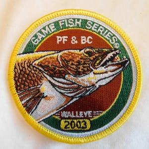 May include: Embroidered patch with a circular design featuring a detailed illustration of a walleye fish. The patch includes the text "GAME FISH SERIES" and "PF & BC" in green and white, with "WALLEYE 2003" at the bottom. The border is yellow.