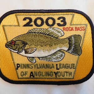 May include: Embroidered patch with a yellow background and a black border. The patch features a detailed illustration of a fish, labeled "Rock Bass," and the year "2003." Text at the bottom reads "Pennsylvania League of Angling Youth."