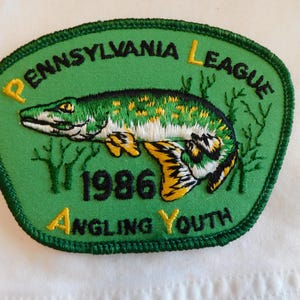 May include: Embroidered patch with a green background and a fish illustration. The patch reads "PENNSYLVANIA LEAGUE ANGLING YOUTH 1986" in yellow text. The fish is green, yellow, and black.