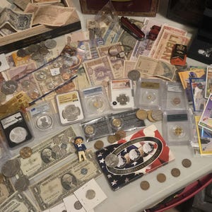 May include: A collection of currency, coins, and collectibles. The image features various paper bills, coins, and graded coins in protective cases. There are also trading cards, a small toy figure, and a jewelry box. The items are arranged on a light-colored surface.
