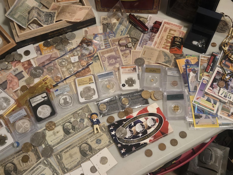 May include: A collection of currency, coins, and collectibles. The image features various paper bills, coins, and graded coins in protective cases. There are also trading cards, a small toy figure, and a jewelry box. The items are arranged on a light-colored surface.