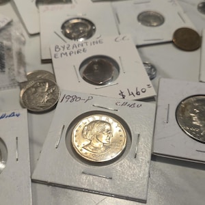 May include: A collection of various silver and gold-toned coins housed in individual white cardboard holders. Several holders feature handwritten text including dates like 1979-D and 1980-P, and the label Byzantine Empire. The coins display portraits and text.