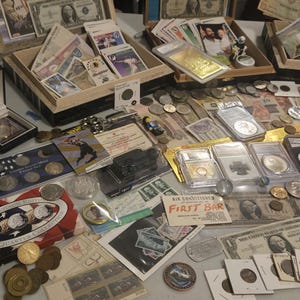 May include: A collection of coins, currency, and collectibles, including US currency, foreign banknotes, and graded coins. Displayed in open wooden boxes and on a table, the items vary in size and shape, with some encased in protective holders.