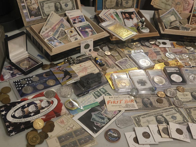 May include: A collection of coins, currency, and collectibles, including US currency, foreign banknotes, and graded coins. Displayed in open wooden boxes and on a table, the items vary in size and shape, with some encased in protective holders.