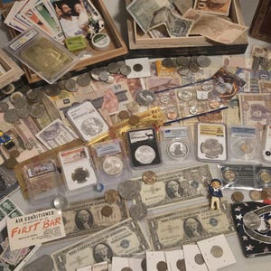 May include: A collection of coins, paper currency, and collectible cards. Several coins are encased in protective holders. There are also various paper bills, including US dollar bills, and a small figurine. The items are arranged on a light-colored surface.