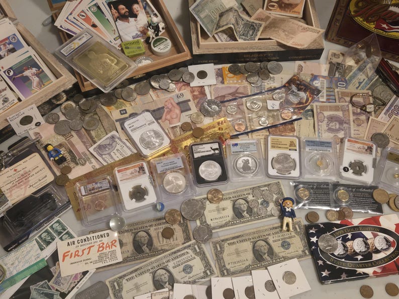 May include: A collection of coins, paper currency, and collectible cards. Several coins are encased in protective holders. There are also various paper bills, including US dollar bills, and a small figurine. The items are arranged on a light-colored surface.
