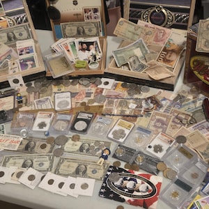 May include: A collection of vintage currency, coins, and collectibles, including paper money, coins, and trading cards. Items are displayed in wooden boxes and on a table, showcasing a variety of historical artifacts.