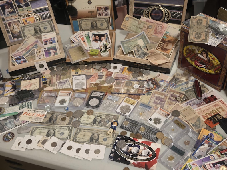 May include: A collection of vintage currency, coins, and collectibles, including paper money, coins, and trading cards. Items are displayed in wooden boxes and on a table, showcasing a variety of historical artifacts.