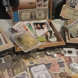 May include: A collection of vintage currency, coins, and trading cards displayed on a table. Includes U.S. dollar bills, foreign banknotes, and a gold bar in a protective case. Various coins and trading cards are also visible.