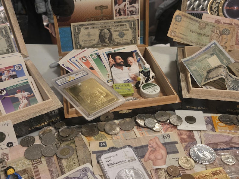 May include: A collection of vintage currency, coins, and trading cards displayed on a table. Includes U.S. dollar bills, foreign banknotes, and a gold bar in a protective case. Various coins and trading cards are also visible.