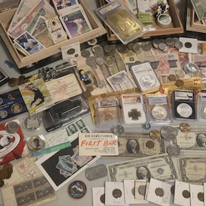May include: A collection of coins, currency, and collectibles, including US coins, paper money, and trading cards. The items are arranged on a table, with some displayed in boxes and protective cases. There are also some figurines.