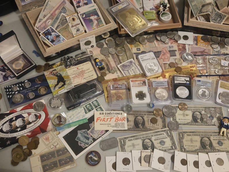 May include: A collection of coins, currency, and collectibles, including US coins, paper money, and trading cards. The items are arranged on a table, with some displayed in boxes and protective cases. There are also some figurines.