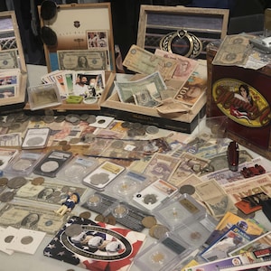 May include: A collection of vintage currency, coins, and collectibles displayed on a table. Items include paper money, coins in protective cases, trading cards, and a decorative cigar box. The items are arranged for sale.